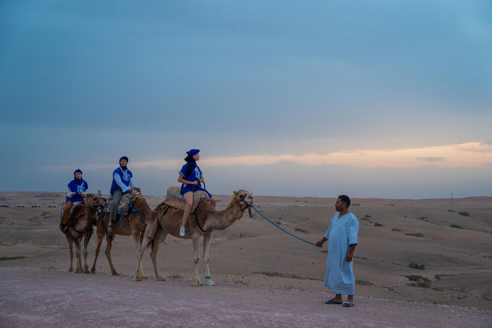 Three riders in blue on camels at dusk, their guide leading them across the plateau