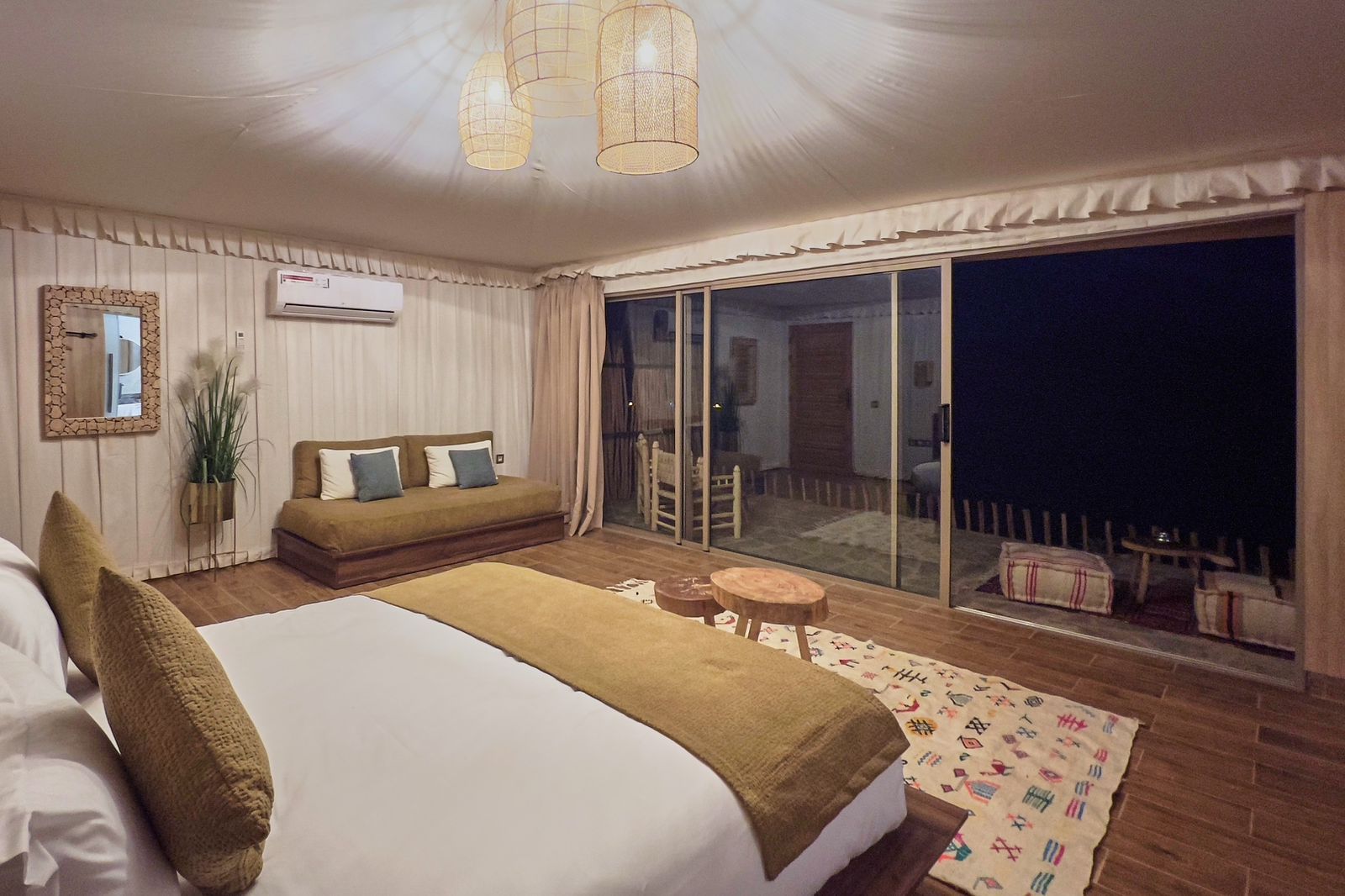 Tent lit in the evening, with a double bed, a day sofa and the dark desert beyond the glass