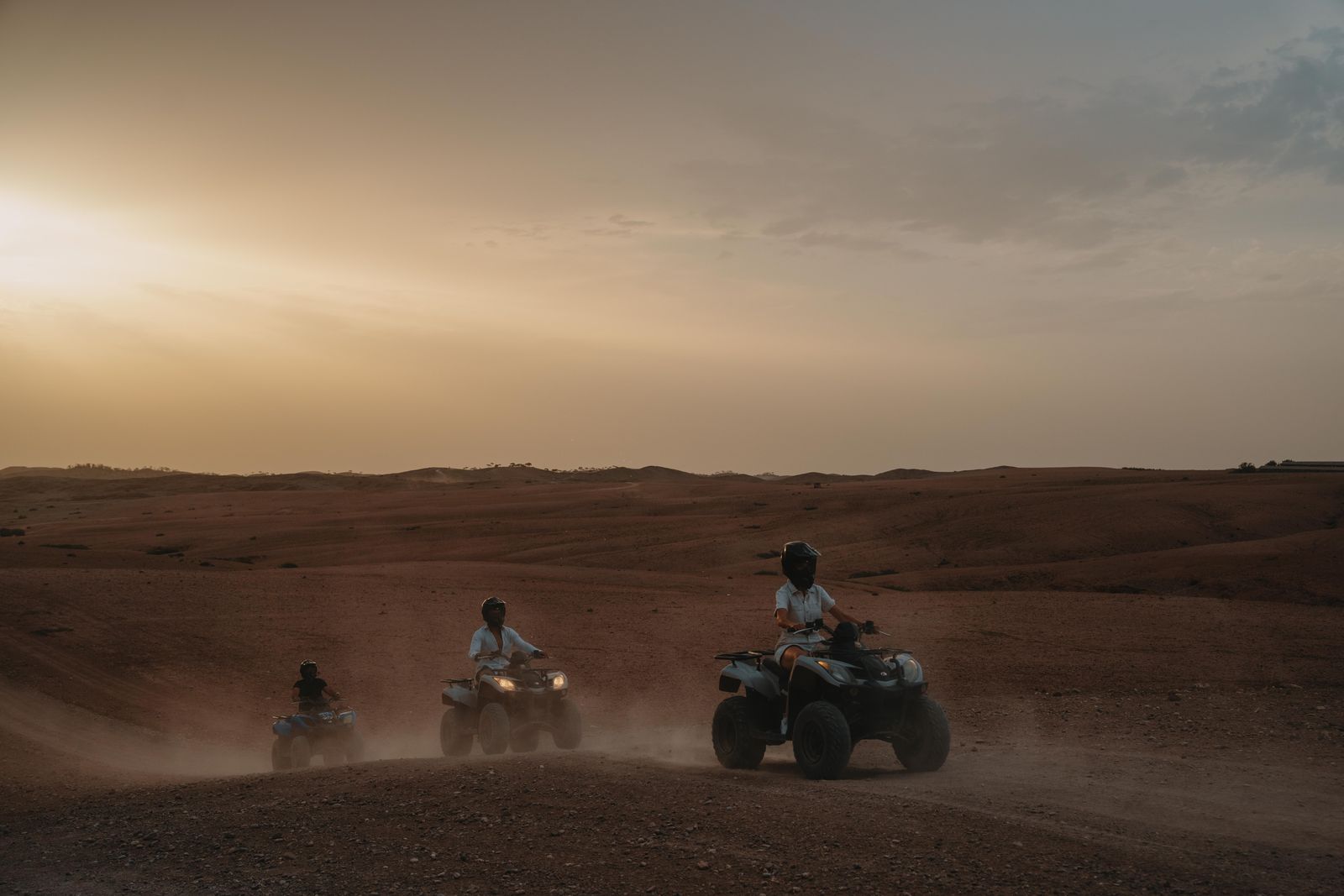Three quad riders on a desert track at sunset, dust rising behind the wheels